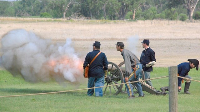 Image of 4 men in 1870s blue dress uniforms, firing a cannon. 