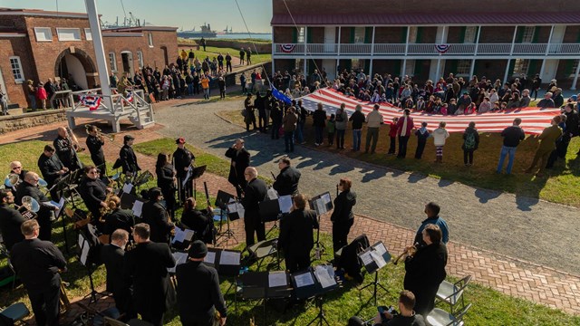 A band and a group of visitors holding a big 15 star 15 stripe flag in the star fort parade ground.