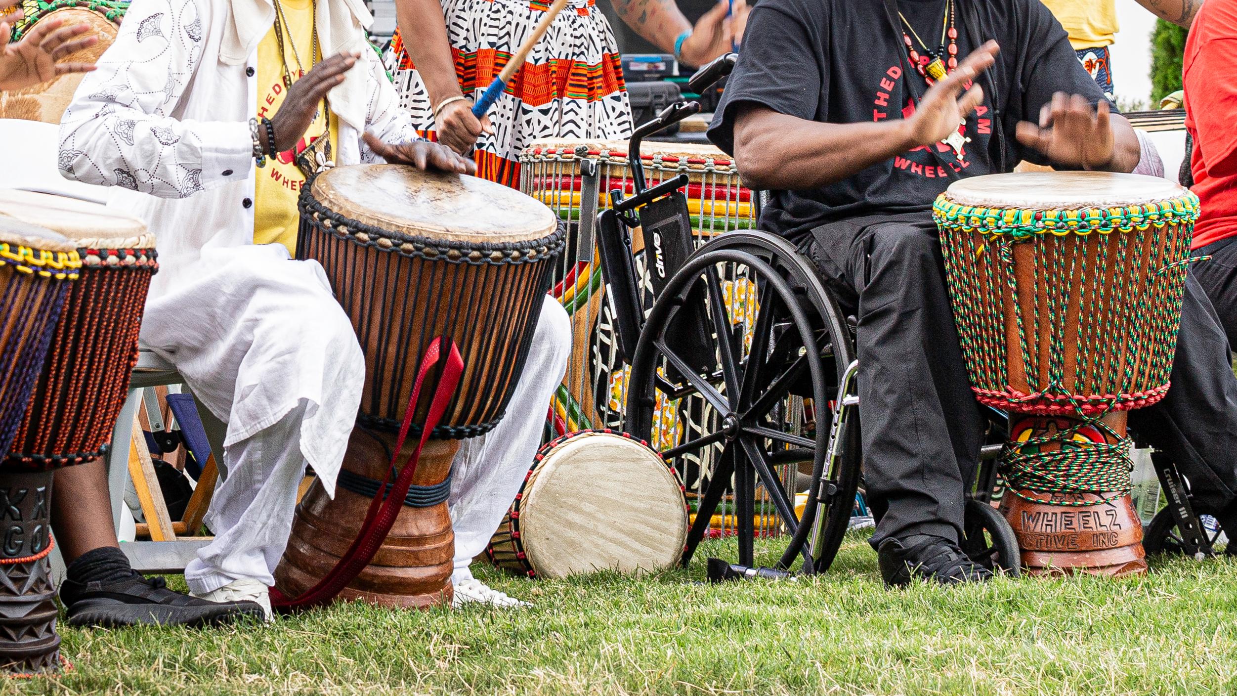 A line of colorfully dressed seated people in colorful clothes beat drums with their hands. .  