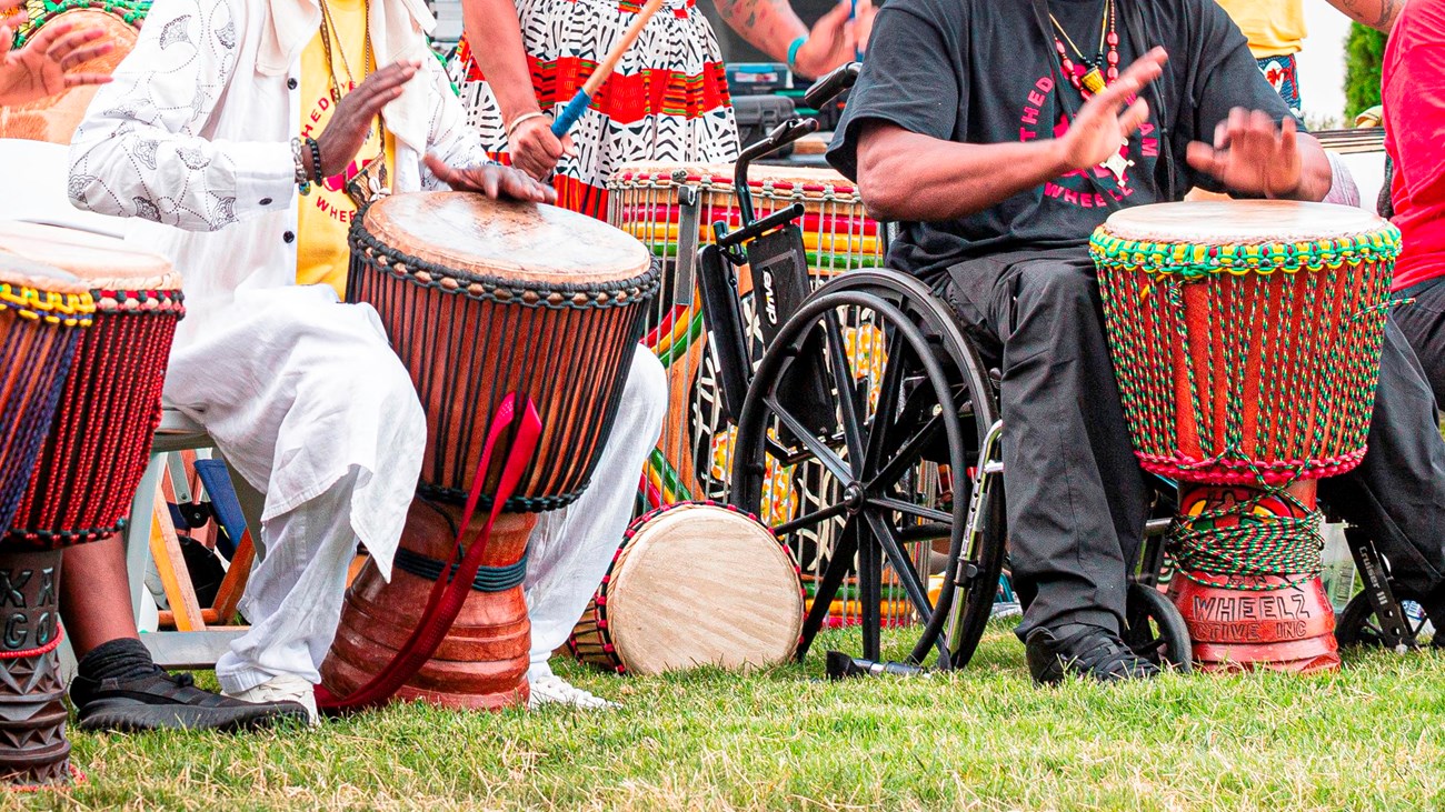 A line of colorfully dressed seated people in colorful clothes beat drums with their hands. .  