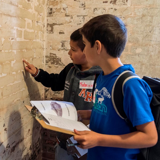 Students stand in front of a brick wall inside Fort Point while holding journals