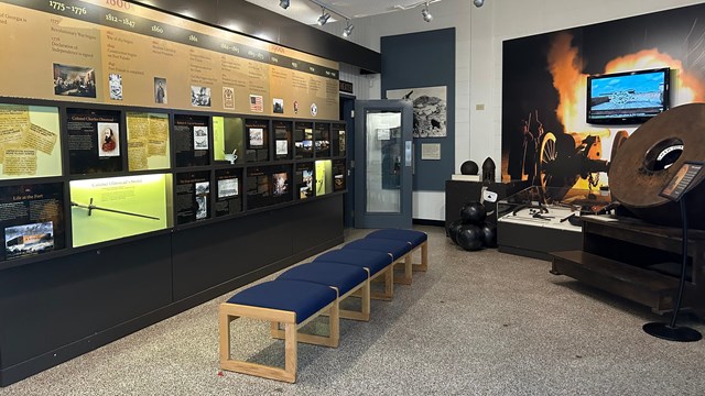 A panel of exhibits on the left, a large cannon and other munitions on the right and center.