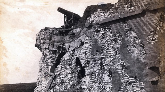 A black and white image of a damaged brick wall with a cannon on top. 