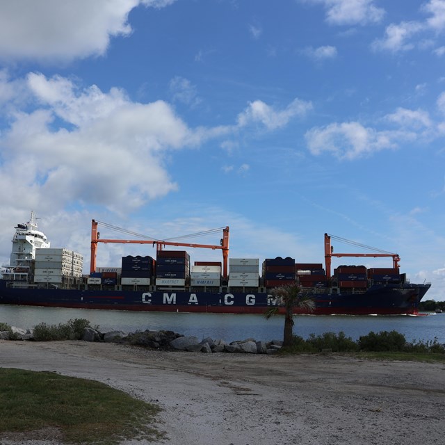 A cargo ship on a river, with a dirt parking area on land. 