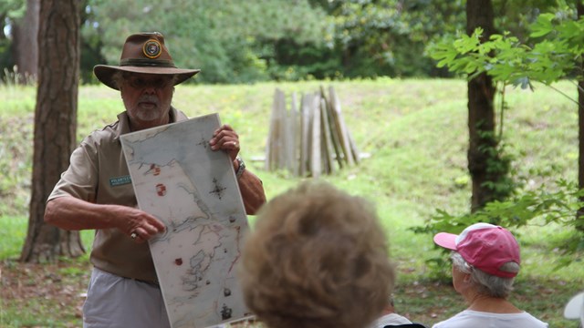 An adult man hold a map pointing at one location in front of a couple adults