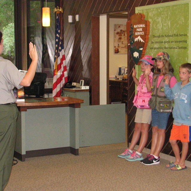color photo of a ranger standing left swearing in three Junior Rangers standing right