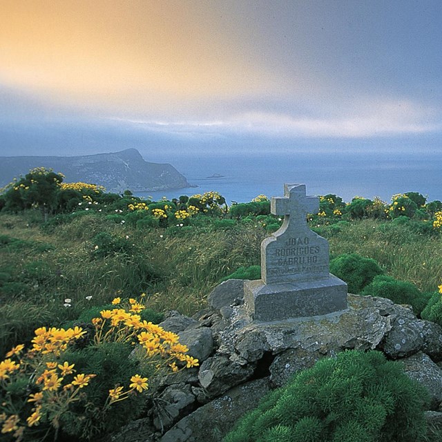a stone marker with cross on wildflower covered hillside overlooking the ocean and islands