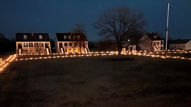 Image of the fort's parade Ground at night lit by candle lanterns. White buildings in the background