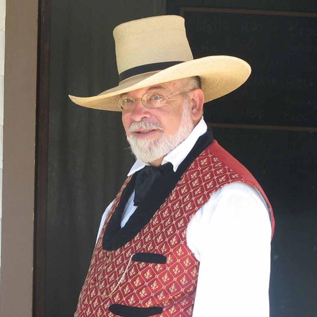 The sutler stands in front of building wearing a straw hat and colorful vest over a white shirt.