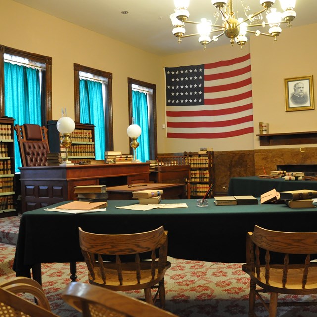 A courtroom, judge's bench rising above tables with green cloths and chairs, a US flag on the wall.