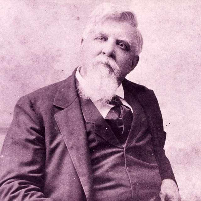 In black and white, an older man with a white beard sits in a chair wearing a dark three piece suit.