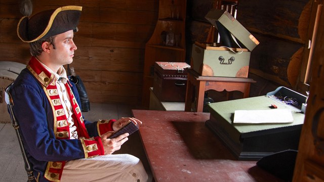 A man sits with with a sheet of paper, a quill pen and ink, and a flute of wine. 