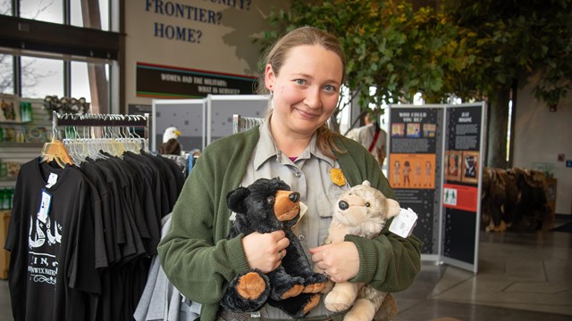 A park ranger snuggles an bear and a wolf stuffie while she smiles contently.