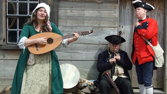 A woman and two men sing together. The woman holds an old oval-shaped guitar. 