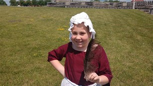 A young girl smiles at the camera. She is wearing an 18th C dress and standing in field of grass.