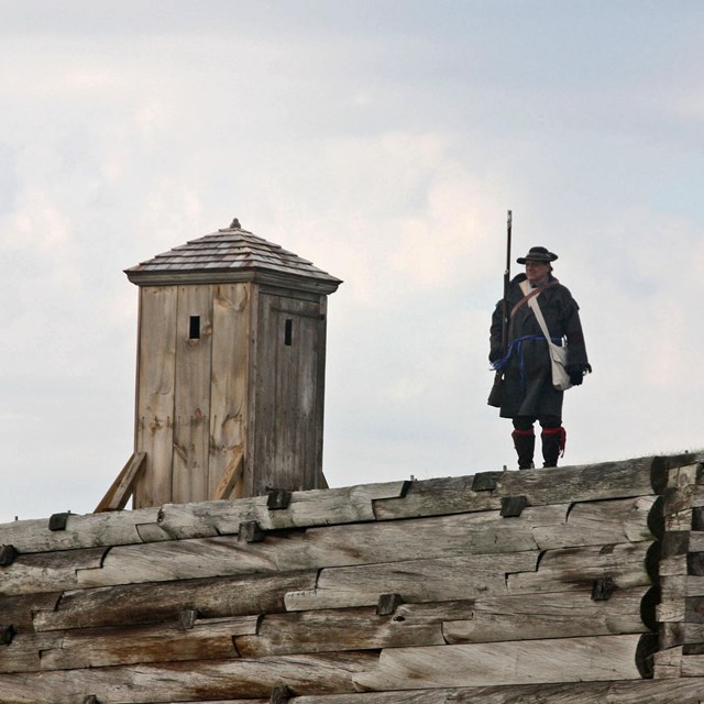 A man in a heavy wool jacket stands with his musket near a sentry box on the fort wall. 
