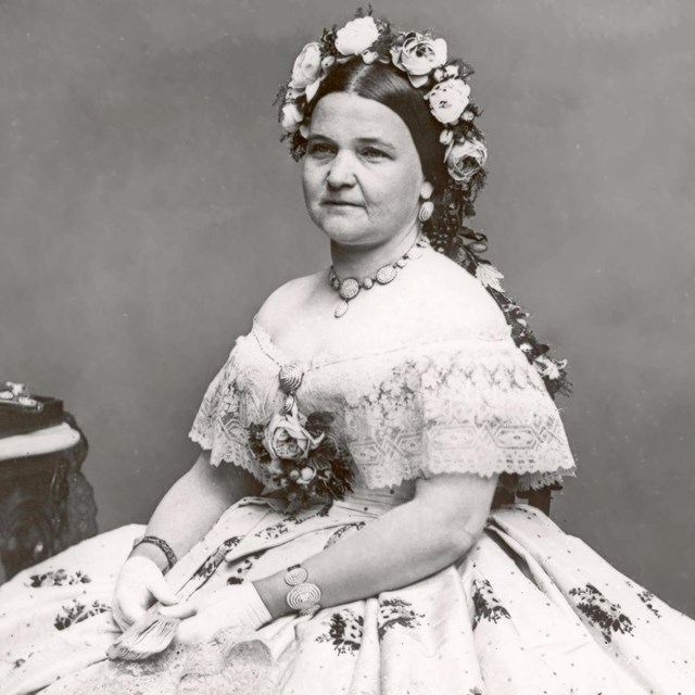 Photograph of a woman in a white dress with flowers in her hair sitting in a chair.