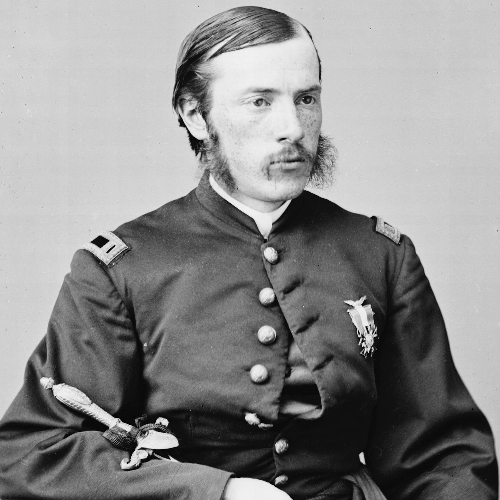 A man with a mustache, wearing a blue Union soldier's coat, and holding a sword sits in a chair