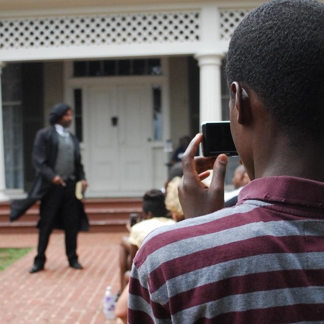 A young man takes a photograph of an actor portraying Frederick Douglass