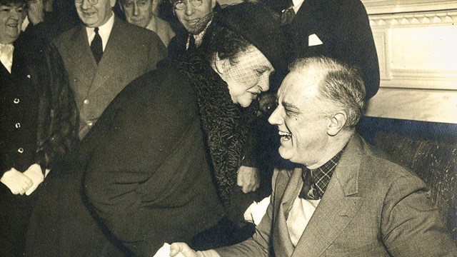 Frances Perkins leans in to shake President Franklin Delano Roosevelt's hand as he smiles