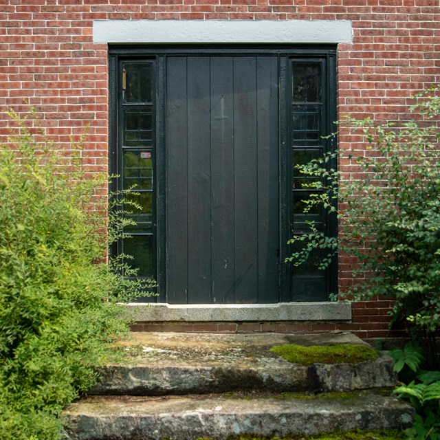 A black wooden door to a red brick building at the top of three mossy stone stairs.