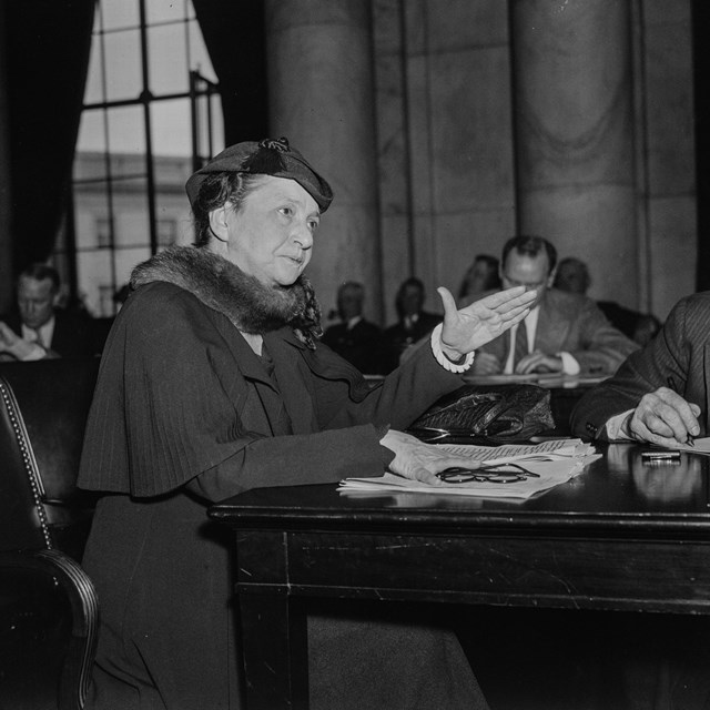 Francis Perkins seated at a desk, left hand raised, palm down, as if making a point in a speech.