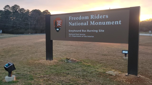 A sign saying Freedom Riders National Monument Greyhound Bus Burning Site is posted in a field.