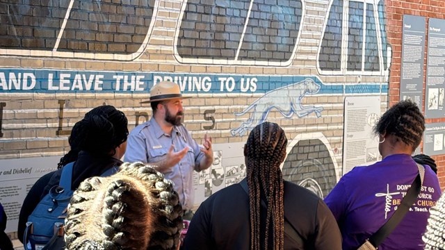 A group of seven people listen to a park ranger talk while standing in front of a bus mural.