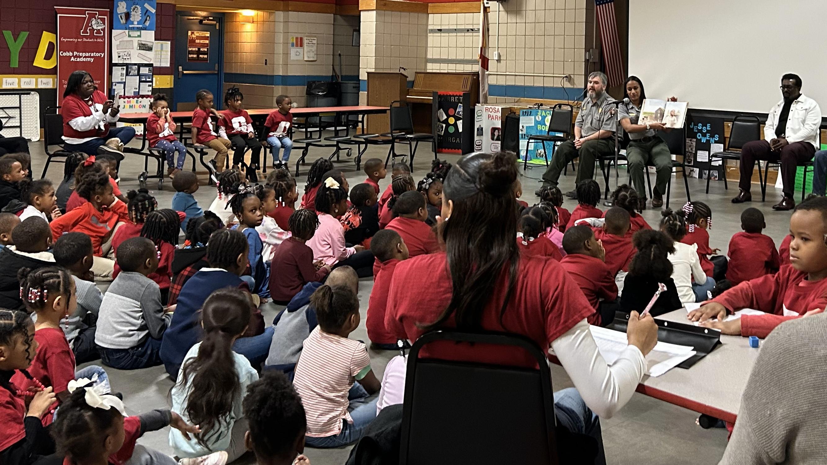A group of about 100 children and teachers listen to rangers reading from a book. 