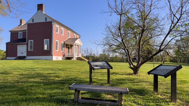 Two interpretive signs in front of a red two story farm house on a field of mowed green grass.