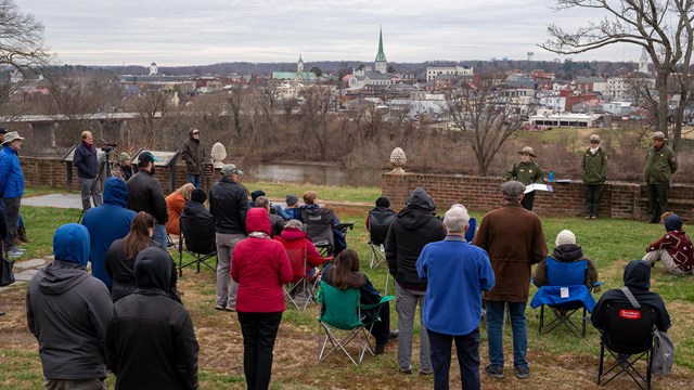 A young female park ranger talks to a group of people by a riverfront with a view of Fredericksburg.