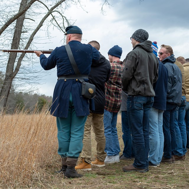 A man in Civil War Union uniform drills a group of military members in civilian clothing in a field.