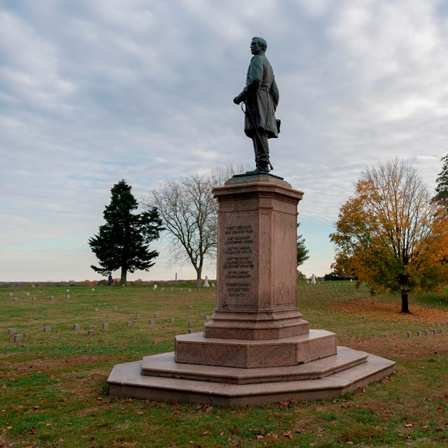 A statue of a man in an open cemetery with fall trees behind.