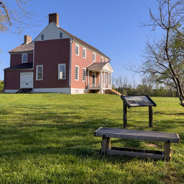 A two-story farm house situated in a field, behind a bench with signs nearby