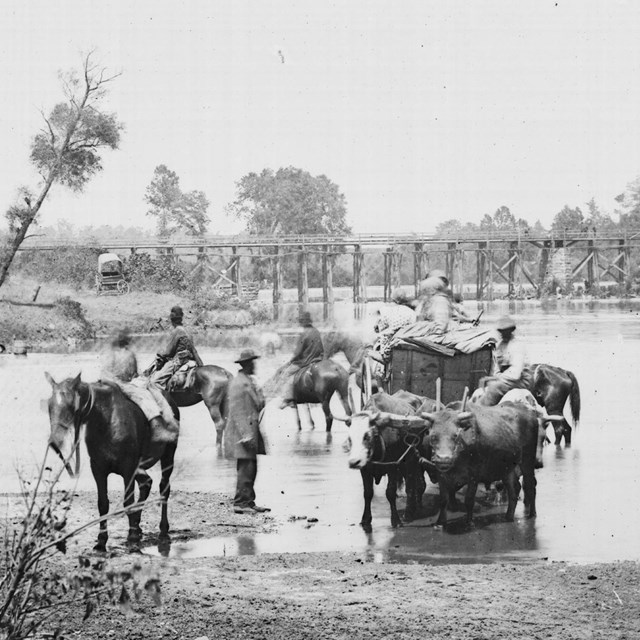 Black and white photograph of Black Americans crossing a low river with wagons and soldiers nearby.
