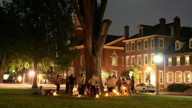 A group of people stand and sit around a tree on grass with luminaries at their feet. 