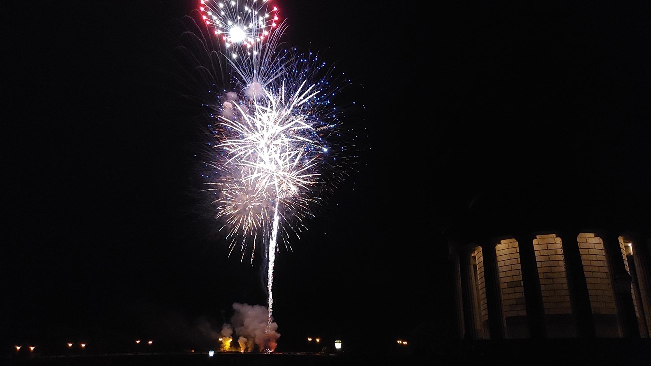 Fireworks in a dark sky next to a the lit Clark memorial