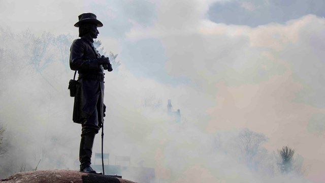 A statue of a soldier is silhouetted against a backdrop of heavy smoke.
