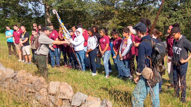 A park ranger leads a group of children on an education program along a stone wall with flags. 