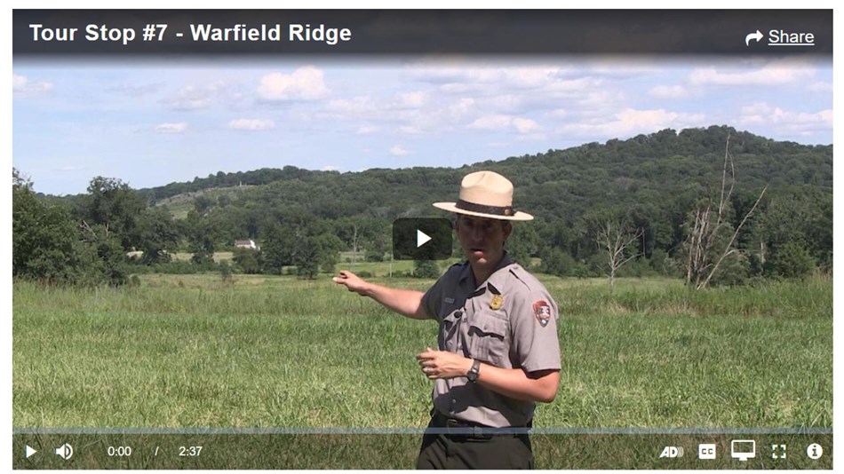 A park ranger points across a green field to two hills in the background. 