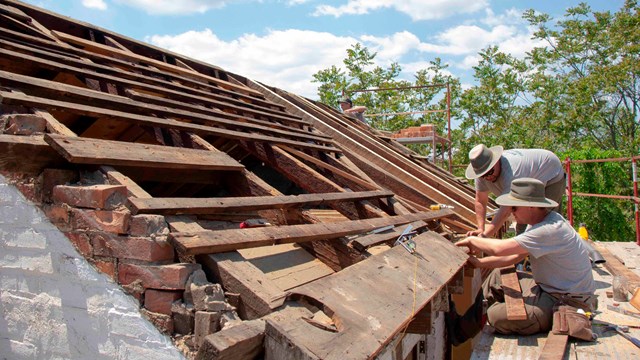 Two workers at the lower right corner assess work on original roof beams on an old house.