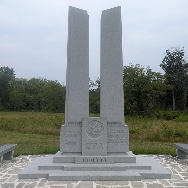 A grey stone monument with two distinct pillars surrounded by flag stone paving.
