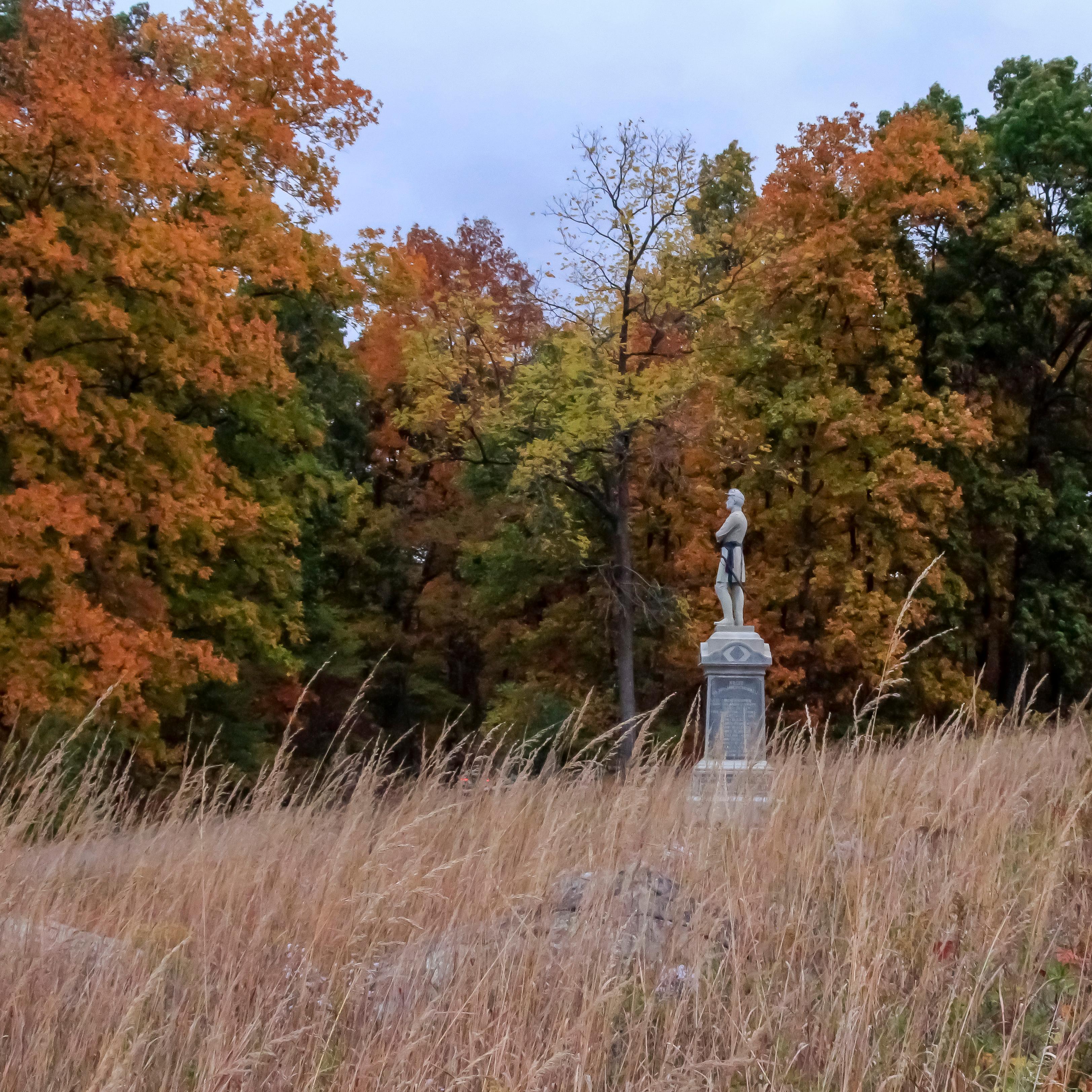 Monument in a field of grass with brightly colored trees behind.