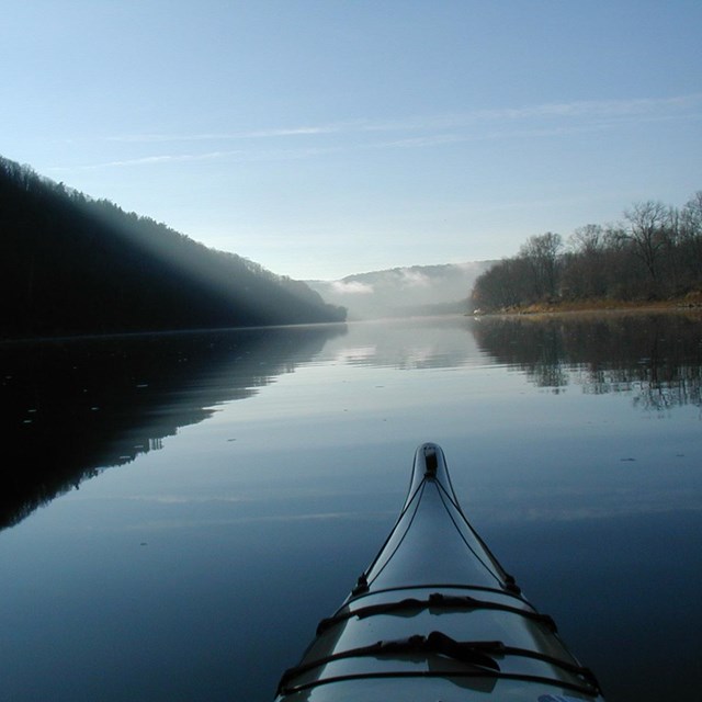 The end of a blue kayak on a body of water with trees and mist in the distance.