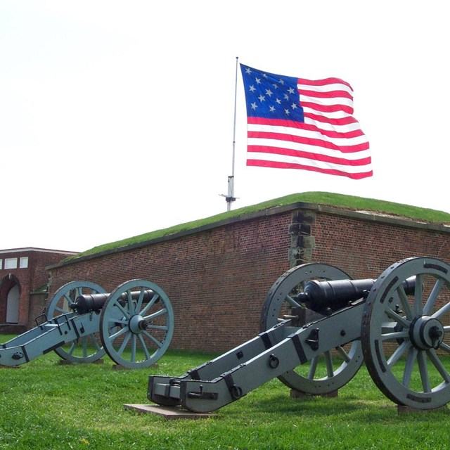 Two cannons sit on grass in front of large red brick fort walls. U.S. flag flys on a flagpole.