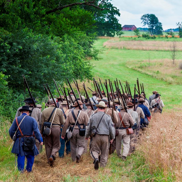 A group of Confederate living historians march across the field. A red barn is in the distance.