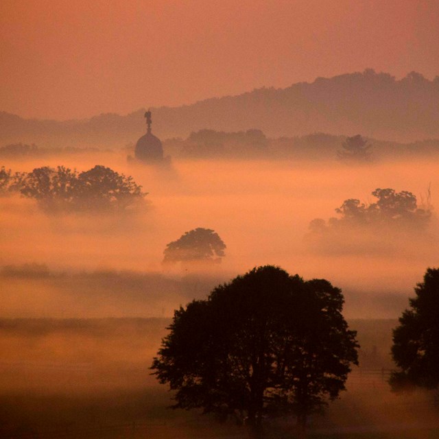 Fog lays heavy on the ground. The sky has an orange glow. Tree tops and a monument are visible.