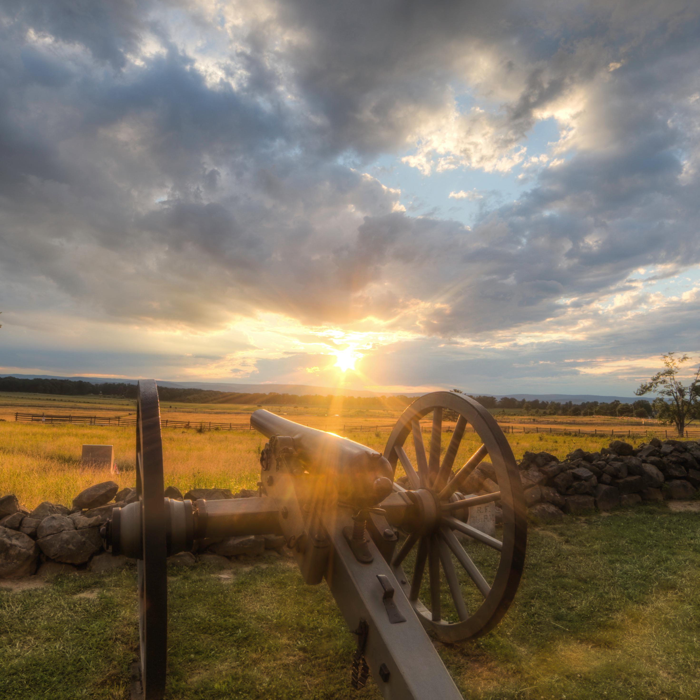 The sun is setting over the battlefield. Two cannons are in the foreground along a stone wall.