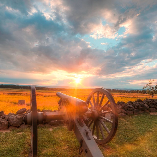 The sun is setting over the battlefield. Two cannons are in the foreground along a stone wall.