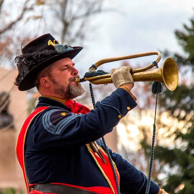A man in a blue uniform and hat is playing the bugle. He wears a red sash over his right shoulder.